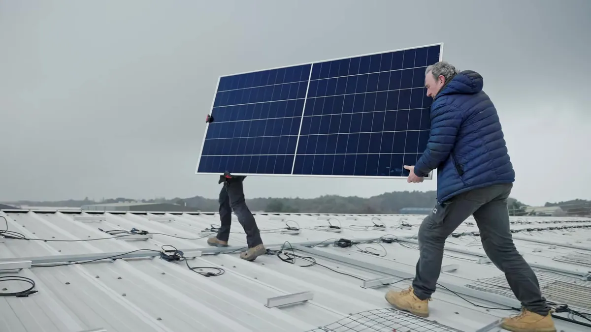 Two engineers carry a large solar panel across a grey industrial rooftop.