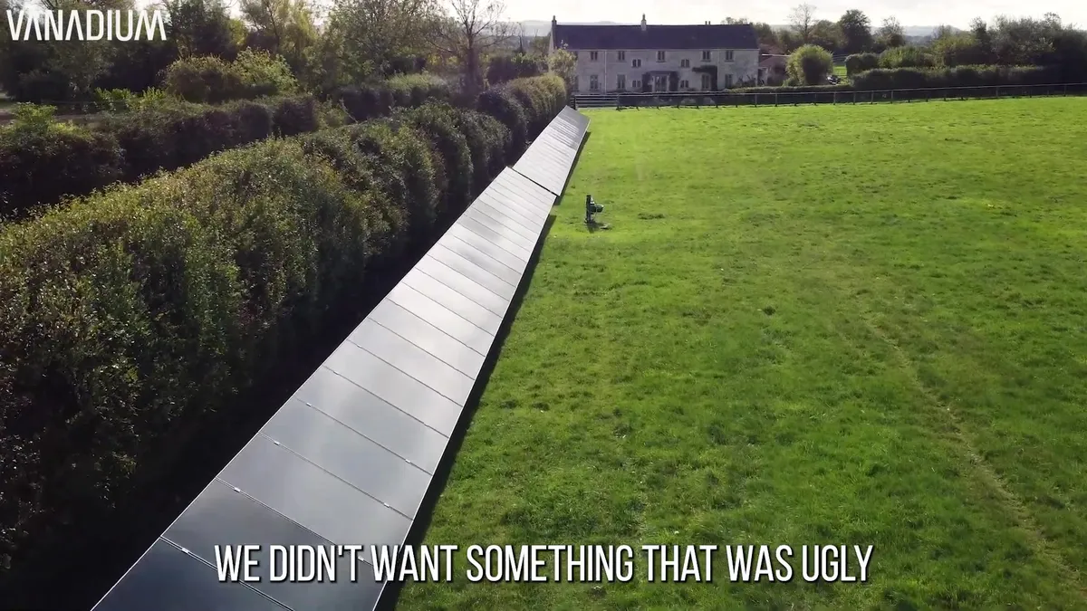 A long row of ground-mounted solar panels runs alongside a hedge and a field, with a house in the background.