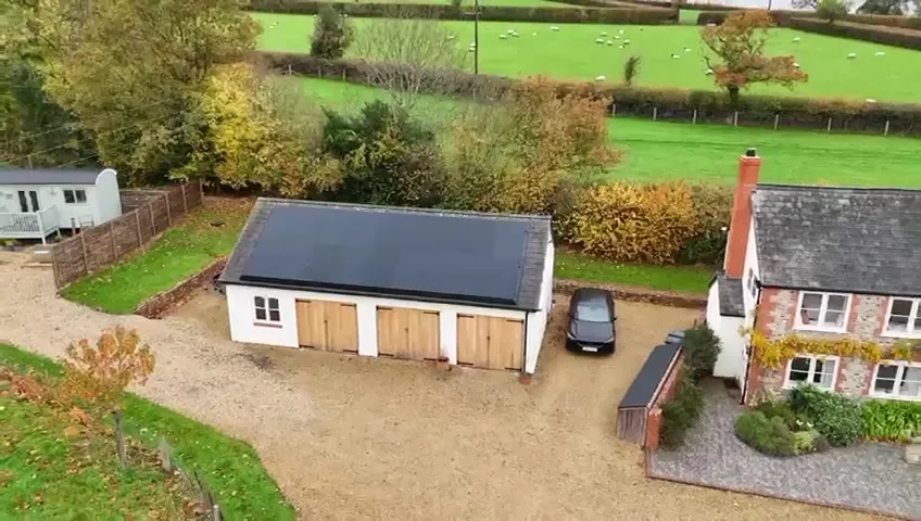 Wide aerial view of the rural property, showing the main house, garage with solar panels, and surrounding green fields.