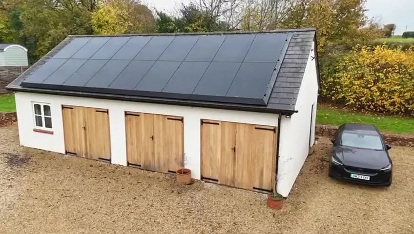 Aerial close-up of the white garage with three wooden doors and 18 black solar panels on its roof.