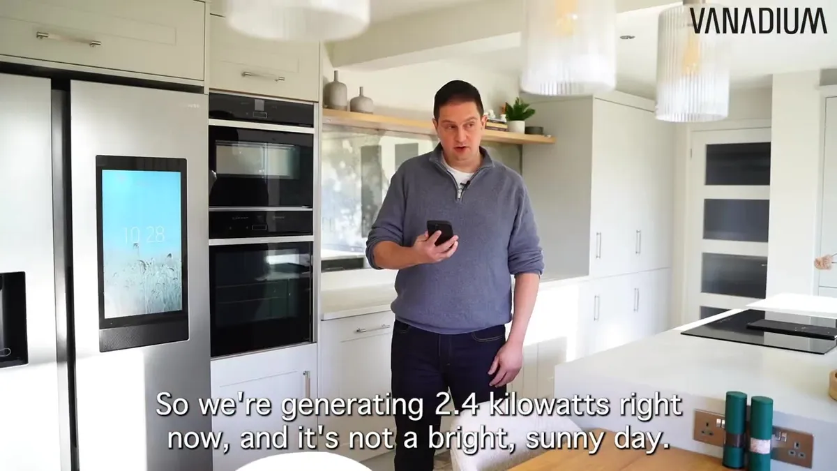 A homeowner in a grey jumper stands in a modern kitchen, looking at his phone with a digital display on a refrigerator to his left.