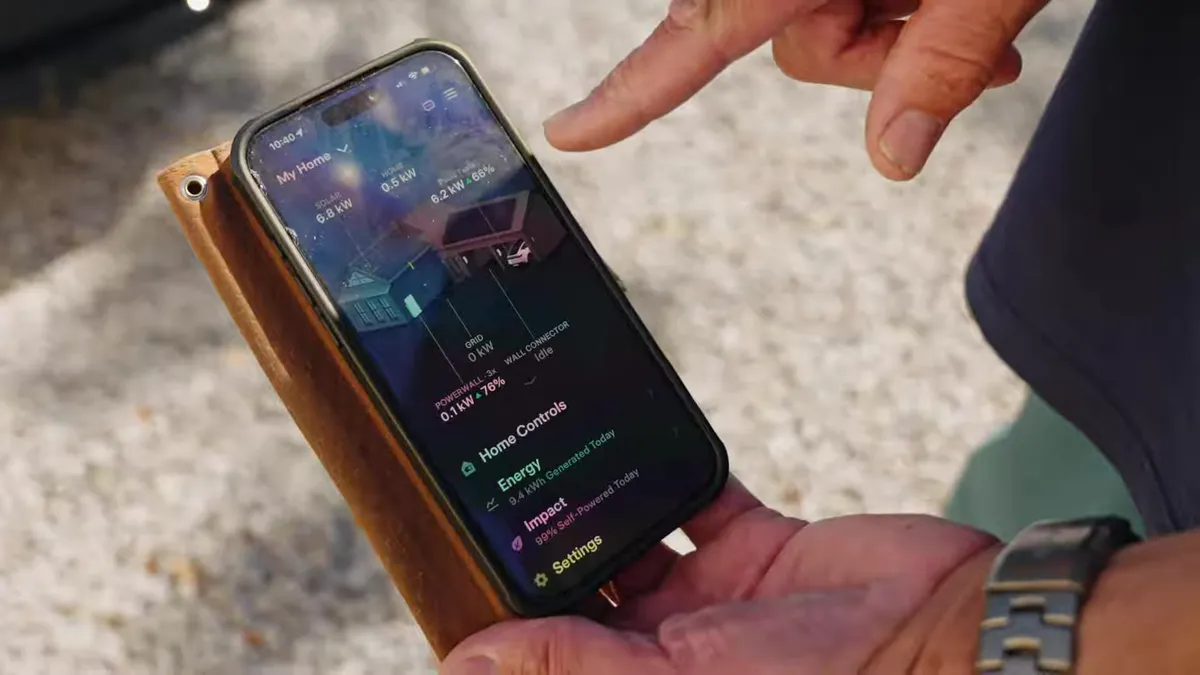 Hand holding a smartphone displaying the Tesla app with home energy usage and Powerwall status.