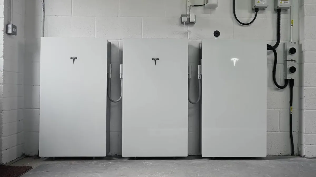 Three white Tesla Powerwall batteries are installed against a white brick wall.