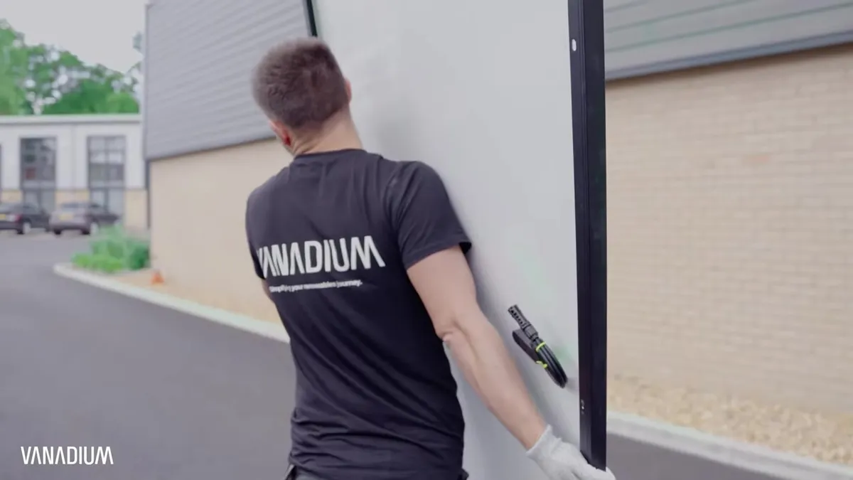 An engineer carries a solar panel past a building on a paved area.